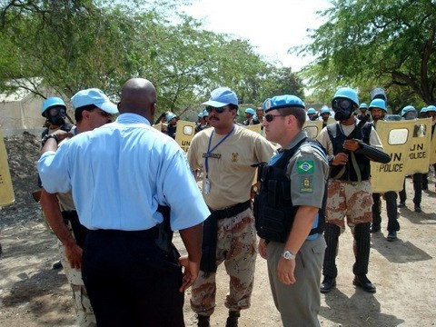 Um homem negro de camisa azul de costas com a mão esquerda sobre um soldado de boné azul conversa com outros 2 integrantes da ONU, em frente a vários integrantes da polícia das nações unidas