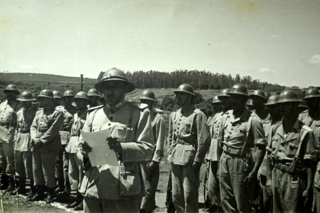 Oficias fardados lendo carta, em homenagem a um coronel morto