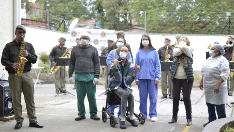 Ao fundo policias militares da Banda de Música da Ajudância-Geral da Brigada Militar com instrumentos musicais e a frente membros da Casa do Excepcional Santa Rita de Cássia posando para a foto.
