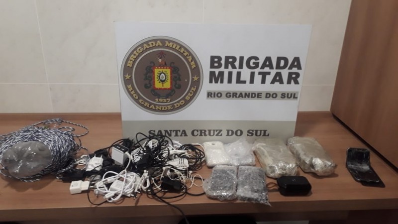 Uma mesa com os materiais apreendidos (Drogas, celulares, entre outros) e no fundo o banner da Brigada Militar