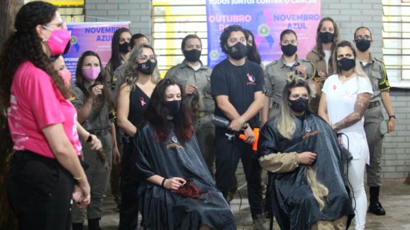 Na frente duas alunas-soldados da Brigada Militar sentadas cortando mechas de cabelo e atrás outras alunas exibindo suas mechas já cortadas.