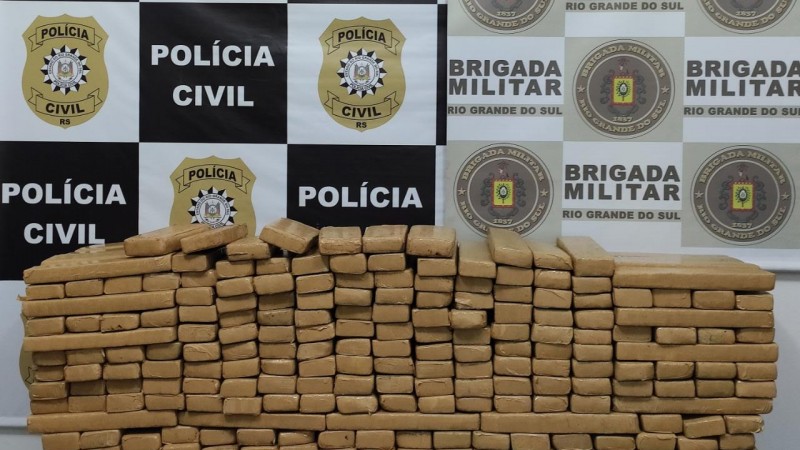 Na imagem uma pilha de tijolos de drogas embaladas. Ao fundo dois banners, um com símbolos da Policia Civil e outro com símbolos da Brigada Militar