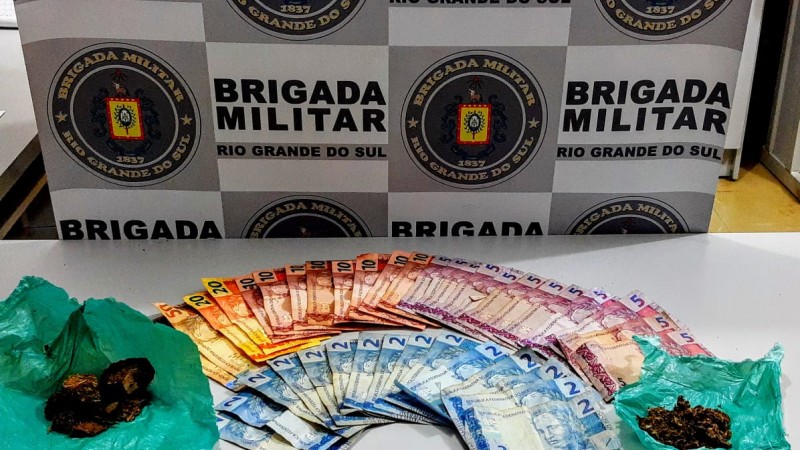 Sobre uma mesa branca drogas e dinheiro em espécie. No fundo o banner da Brigada Militar.