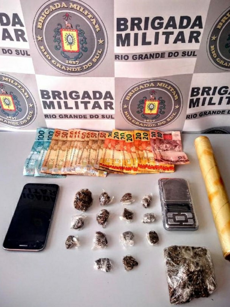 Sobre uma mesa branca esta celular, balança de precisão, cédulas de dinheiro e drogas apreendidas. Ao fundo o banner da brigada Militar.