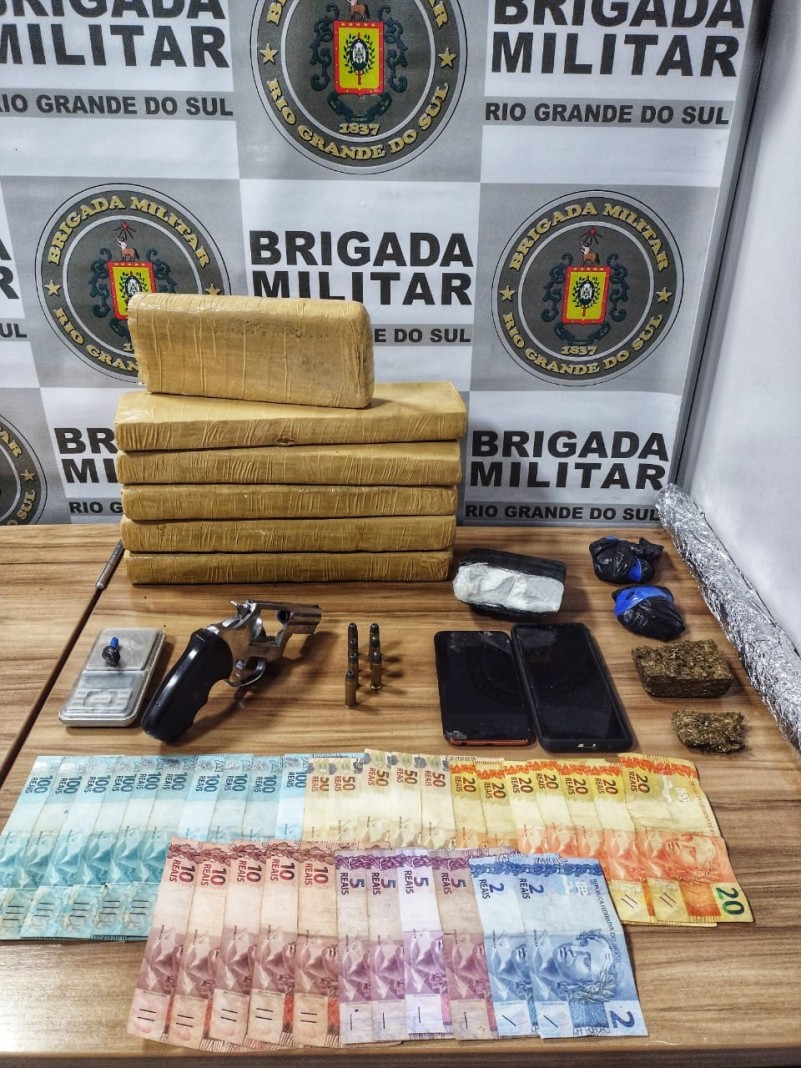 Sobre uma mesa tijolos de drogas e porções fracionadas. Moeda em especie, celulares, uma arma de fogo e munições ao lado. No fundo o banner institucional da Brigada Militar.