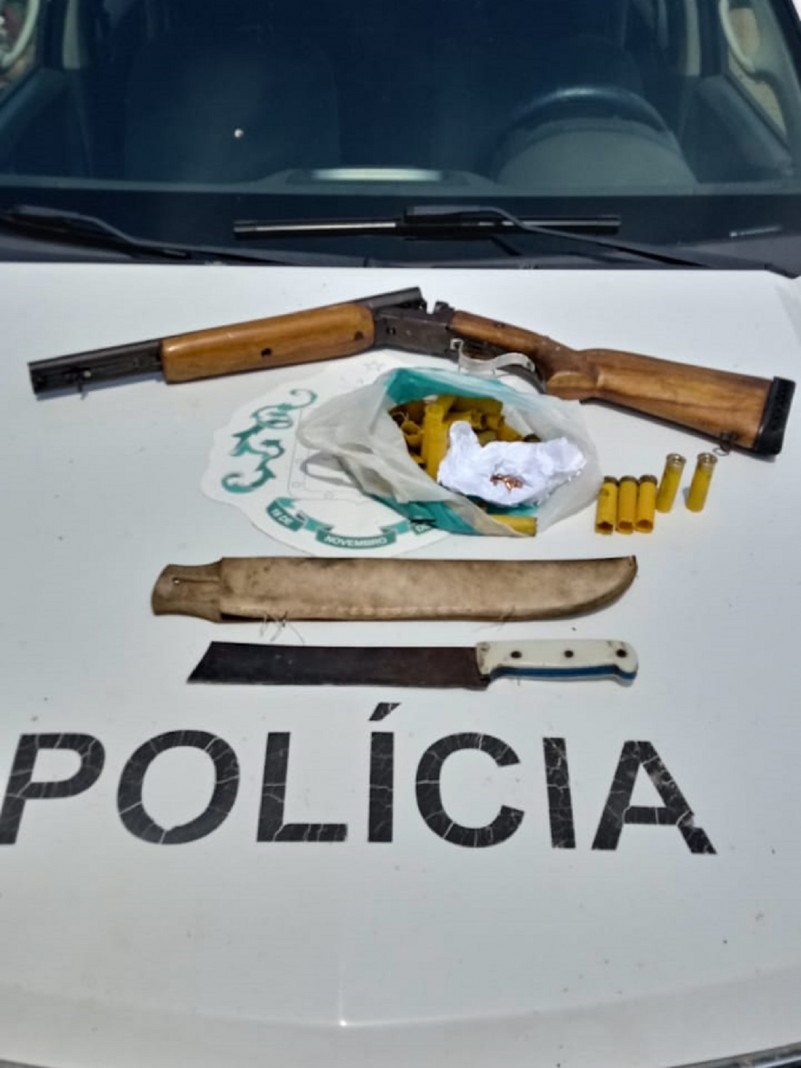 Materiais apreendidos sendo uma espingarda e cartuchos de arma de fogo. Ao lado um facão e uma bainha.