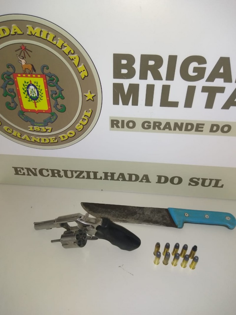 Ao fundo da imagem o banner da Brigada Militar. Sobre uma mesa uma arma de fogo, munições e uma faca.
