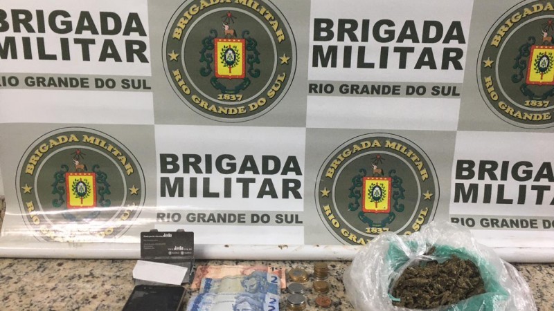 Ao fundo o banner da Brigada Militar. Sobre uma mesa drogas, moeda em espécie e um celular.
