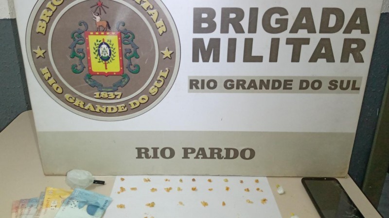 No fundo o banner da Brigada Militar. Sobre uma mesa drogas, celular e moeda em espécie.