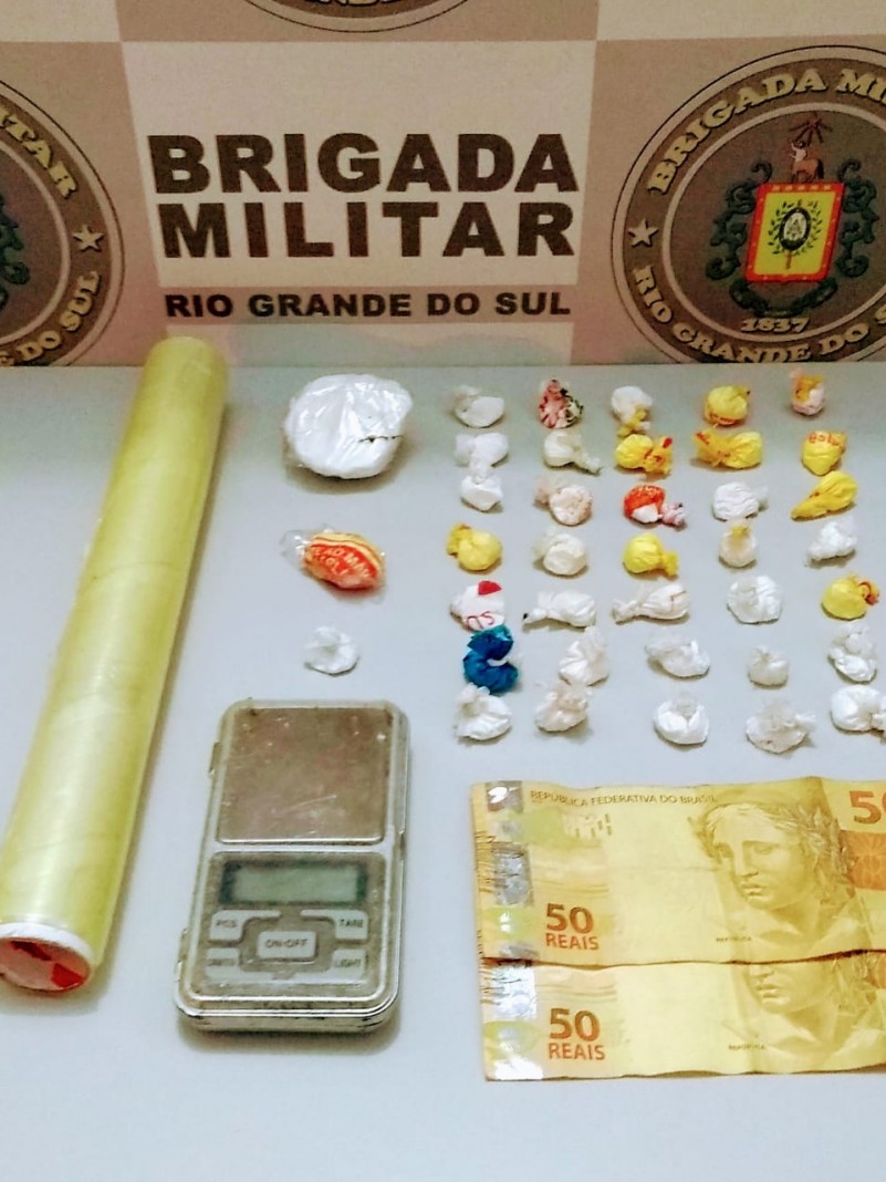 No fundo o banner da Brigada Militar. Sobre uma mesa drogas fracionadas, moeda em espécie e uma balança de precisão.