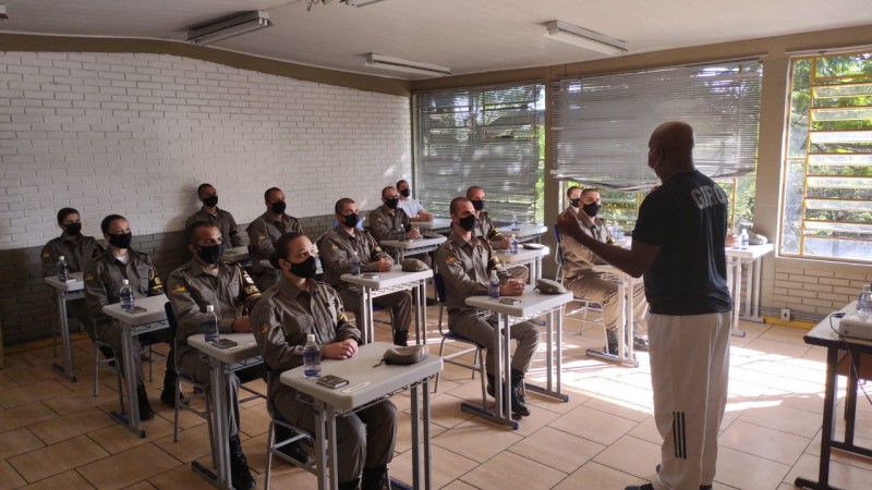 Na foto o professor em frente aos alunos-soldados fardados, os quais estão sentados em cadeiras e mesas escolares dentro de uma sala de aula