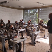 Na foto o professor em frente aos alunos-soldados fardados, os quais estão sentados em cadeiras e mesas escolares dentro de uma sala de aula