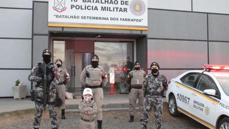 Foto da criança com 5 policiais militares em frente a sede do batalhão. Ao lado uma viatura. 