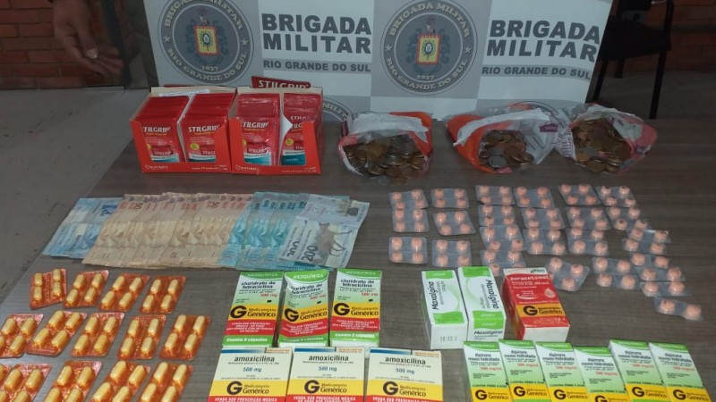 Ao fundo o banner da Brigada Militar. Sobre uma mesa diversas caixas de remédios e comprimidos avulsos, bem como moeda em espécie