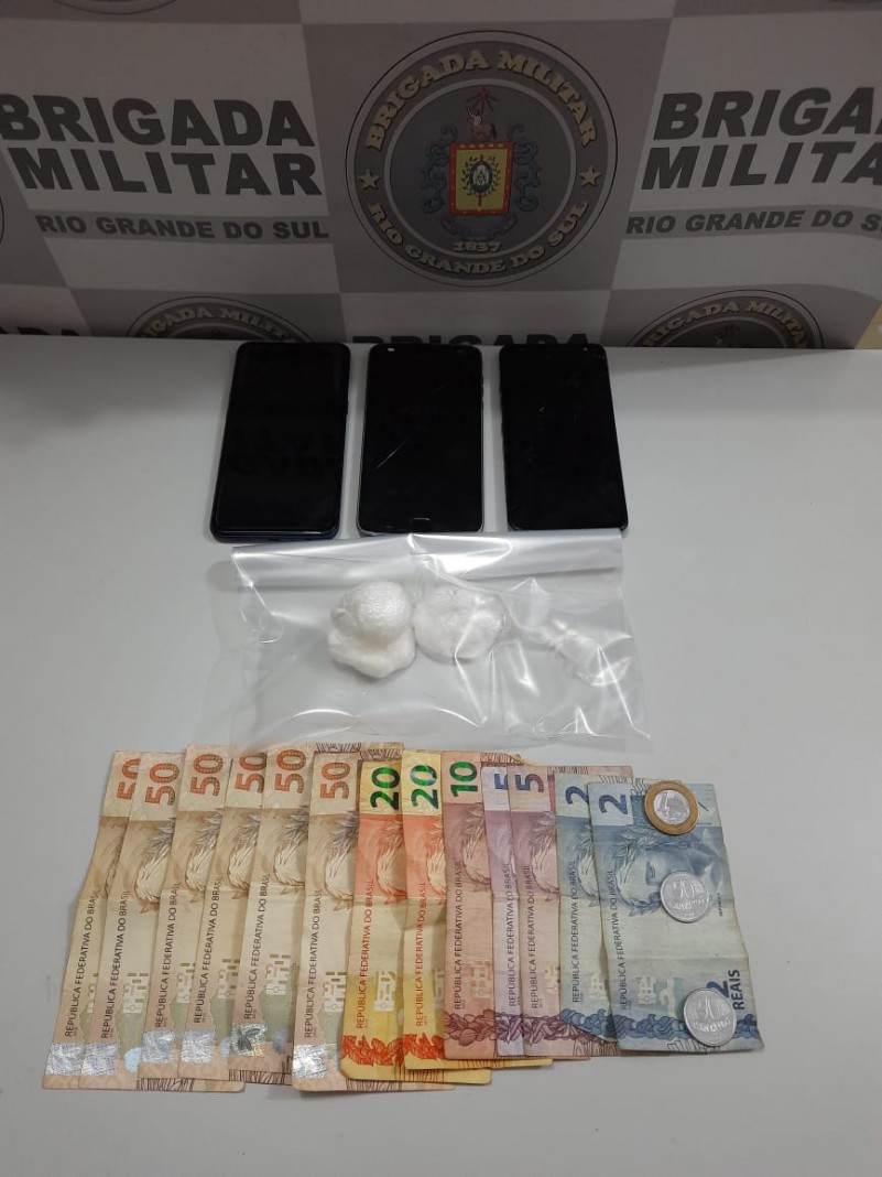 Ao fundo o banner da Brigada Militar. Sobre uma mesa três celulares, drogas fracionadas e dinheiro em espécie.