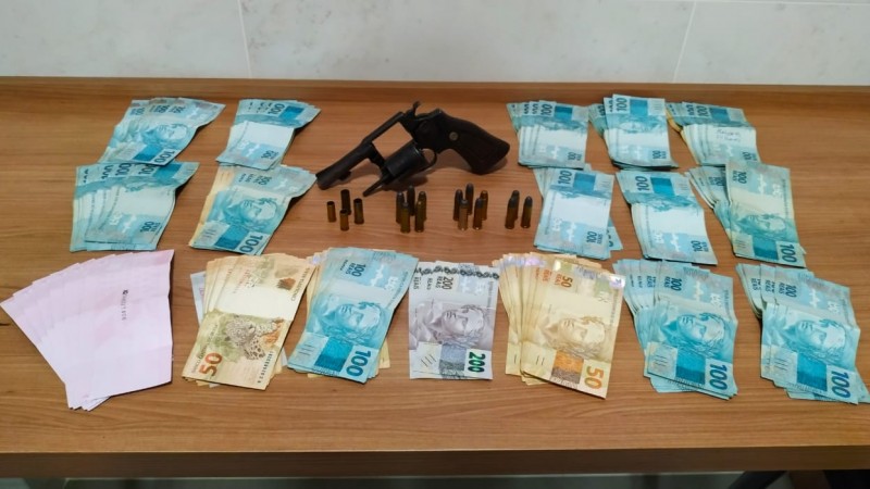 Sobre uma mesa dinheiro em espécie, uma arma de fogo e munições ao lado.