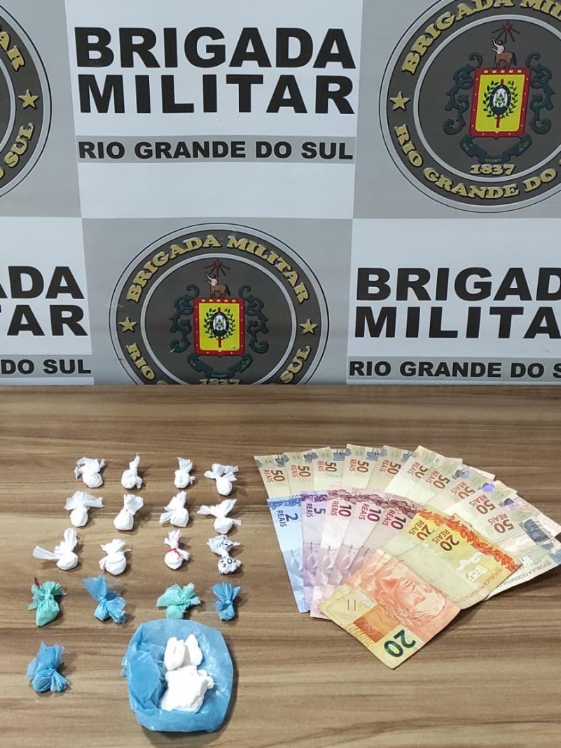 Ao fundo o banner da Brigada Militar. Na frente uma mesa com drogas fracionadas e dinheiro em espécie.