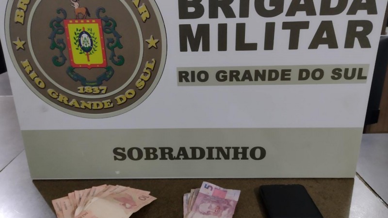 Ao fundo o banner da Brigada Militar. Na frente sobre uma mesa dinheiro em espécie e um telefone celular.
