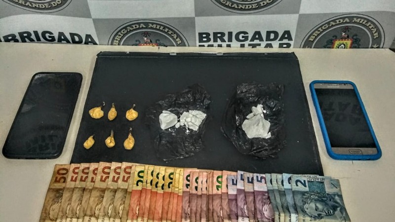 Ao fundo o banner da Brigada Militar, e sobre uma mesa drogas fracionadas, dois celulares e dinheiro em espécie.