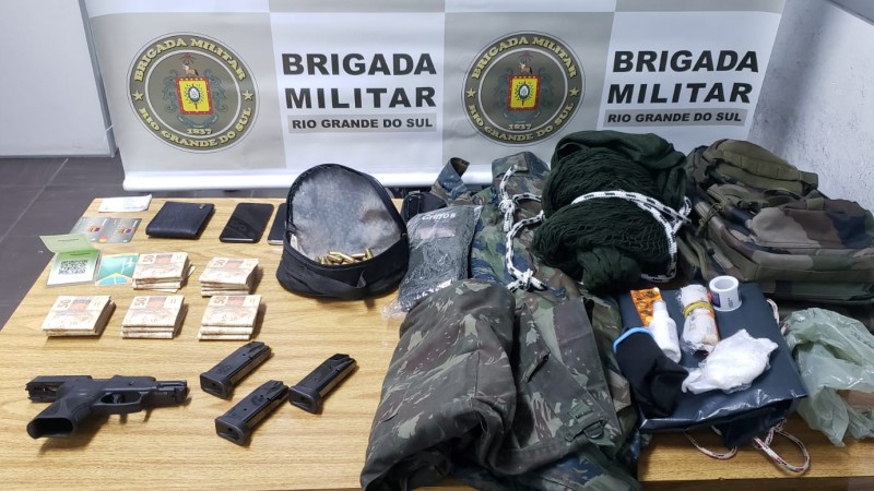 Foto dos materiais apreendidos com o indivíduo. Em cima de uma mesa está a arma, munições, dinheiro, roupas camufladas e outros intens