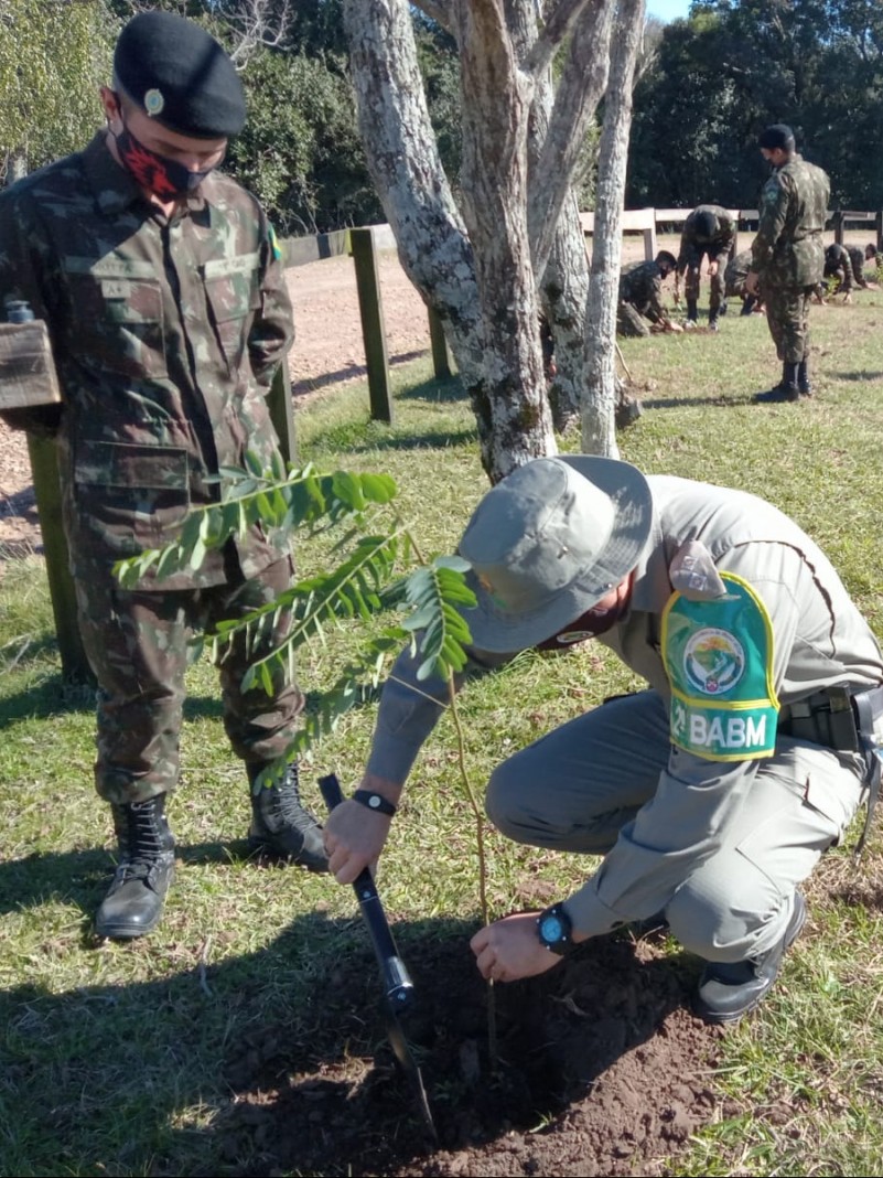 Policial militar ambiental agachado pantando uma muda de árvore. Ao lado, um militar do Exército Brasileiro em pé olhando.