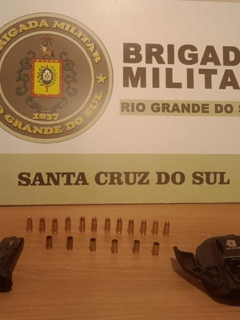 Doze munições intactas e a frente seis estojos de munições deflagradas. Ao lado um carregador e no outro lado um porta carregador. Ao fundo o banner institucional da Brigada Militar de Santa Cruz do Sul.