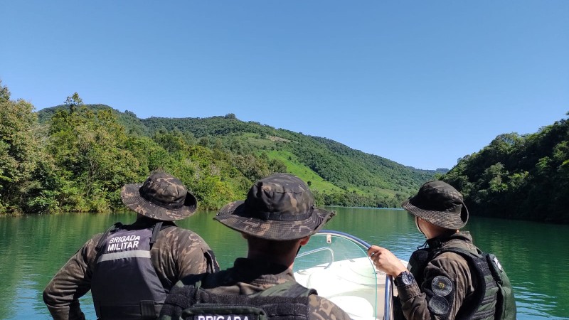 Foto colorida. Três Policiais Militares ambientais vestindo farda camuflada de cor verde escura e coletes salva-vidas de cor preta, estão de costas , em pé em cima de um barco branco. Navegam em um rio com águas verde turquesa e estão rodeados por uma mata verde. O céu é azul e não há nuvens