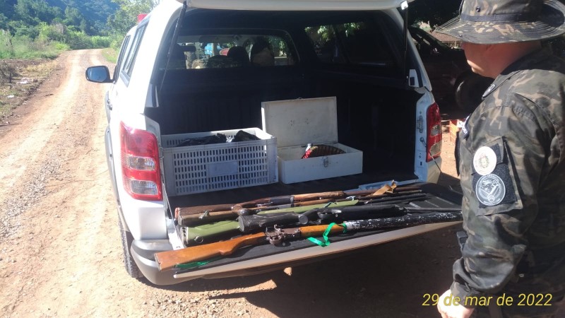 Foto colorida. No primeiro plano, canto direito da foto, um Policial Militar Ambiental, vestindo farda camuflada em tom de verde escuro e usando um chapéu de tecido camuflado, observa a traseira de uma viatura policial, modelo caminhonete, com o porta-malas aberto. No porta-malas há seis espingardas e ao fundo duas caixas brancas. A viatura está parada em uma estrada . Ao fundo aparecem árvores verdes em um mato
