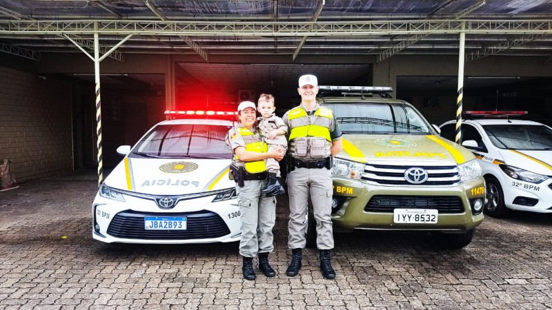 Policial feminina com menino nos braços e Policial Militar masculino postados para foto em frente a viaturas da Brigada Militar