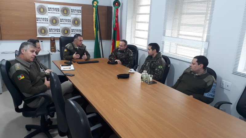 Comandante do CRPO/VRP e Comandante do 7º BIB, juntamente com Oficiais dos Comandos sentados em uma mesa. Ao fundo o banner da Brigada Militar.