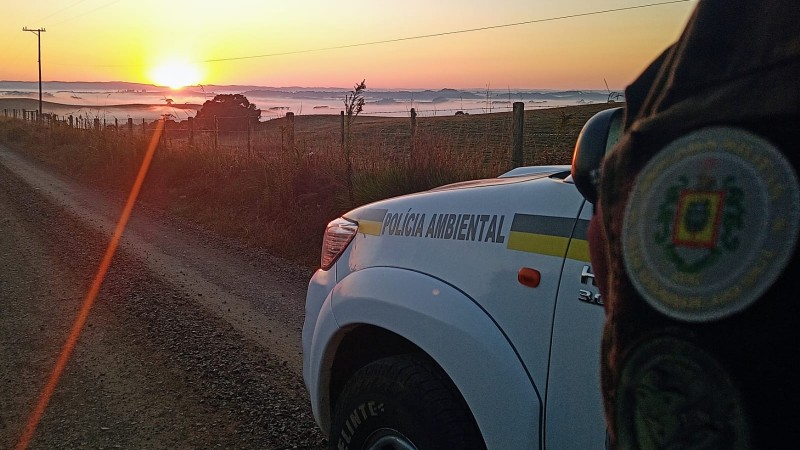 Foto Colorida. Ao fundo, o nascer do sol no horizonte. No canto direito da foto uma viatura policial , branca com listras amarela e verde milita. Posicionado ao lado da viatura um policial, em pé, com fardamento camuflado em tons de verde escuro. A frente da viatura uma estrada e ao lado um campo - tipo pastagem.