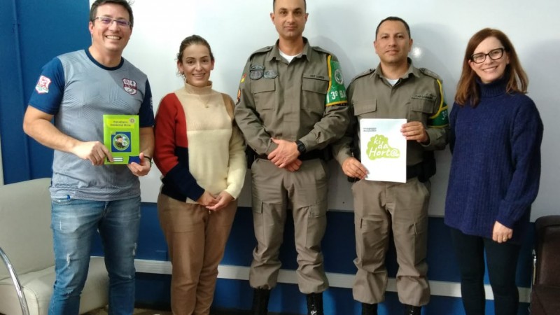 Foto colorida, numa sala com quadro branco e paredes azul. Postados à frente do quadro, dois Policiais Militares e três pessoas civis. Da Esquerda pra direita> um homem branco, alto, veste calça jeans e camiseta cinza com mangas azuis, esse segura uma Cartilha do Programa Patrulheiro Ambiental Mirim de cor verde; ao lado uma mulher, branca, baixa, vestindo calça caramelo e blusa manga longa bege com detalhes em vermelho e marinho; ao lado um Policial militar, branco, alto , vestindo farda de cor cinza e um bracelete verde; à direita, outro policial militar, homem, branco, mais baixo e vestindo farda cinza com bracelete verde, esse policial segura uma Cartilha Branca; ao seu lado uma mulher mais baixa, branca e ruiva, vestindo calça preta e blusão de lã azul marinho.