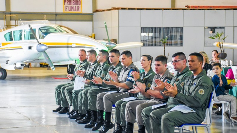 Imagem mostra os alunos formados sentados aplaudindo e, em segundo plano, um avião turbo-hélice 