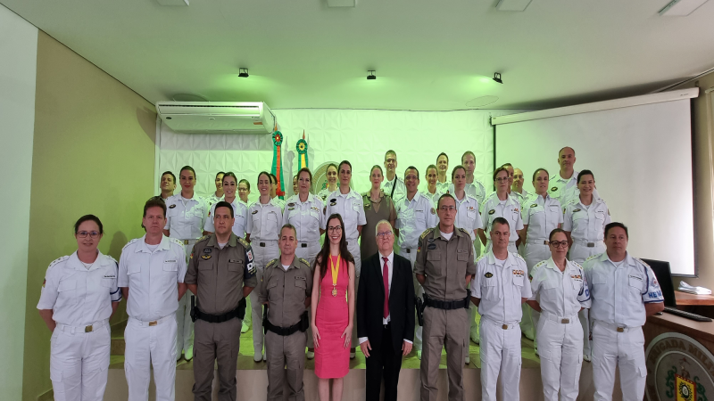 Grupo de pessoas com diferentes fardas estão reunidas lado à lado olhando para a câmera, atrás delas a uma sala com luzes verdes.