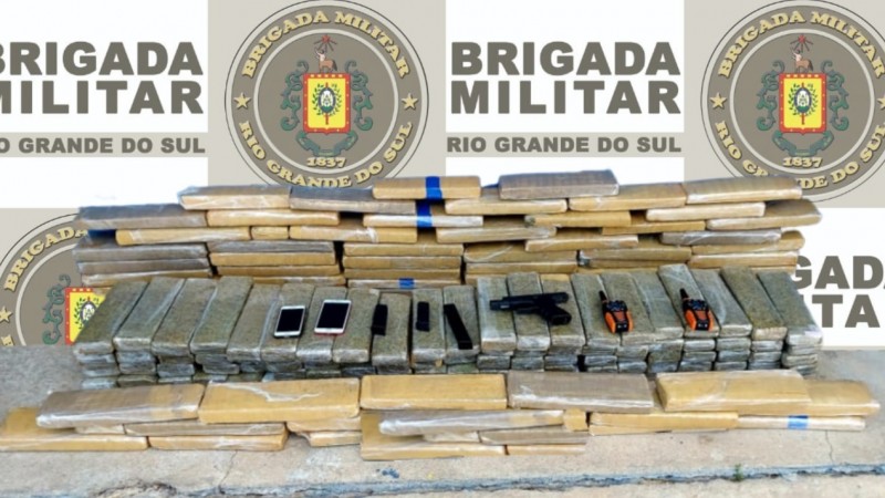 Imagem mostra diversos tabletes de maconha empilhados, com armas, carregadores e celulares em primeiro plano. Em segundo plano, banner institucional da Brigada Militar
