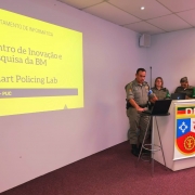 Três militares estão de pé fazendo a apresentação ao lado de uma tela projetada.