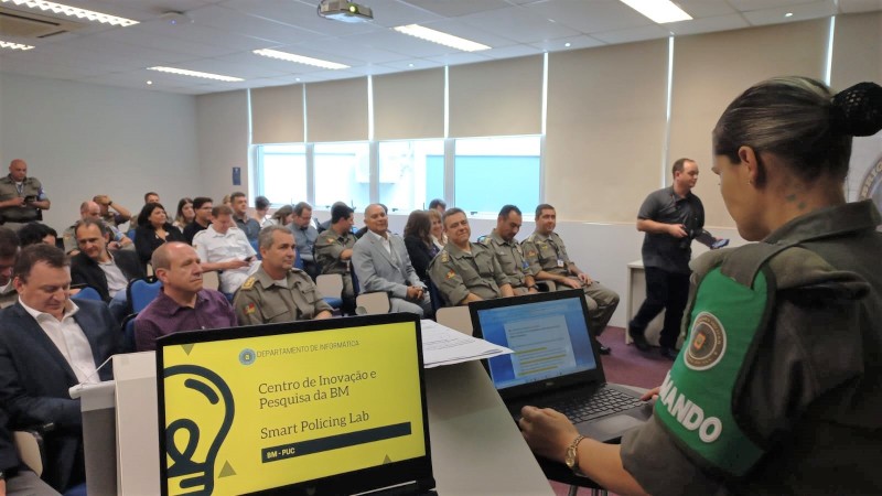Uma militar est de pé ao lado de um notebook, em frente a uma plateia sentada.