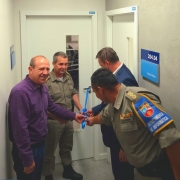 Quatro homens, dois militares e dois civis estão segurando uma fita azul, em frente a uma porta branca.
