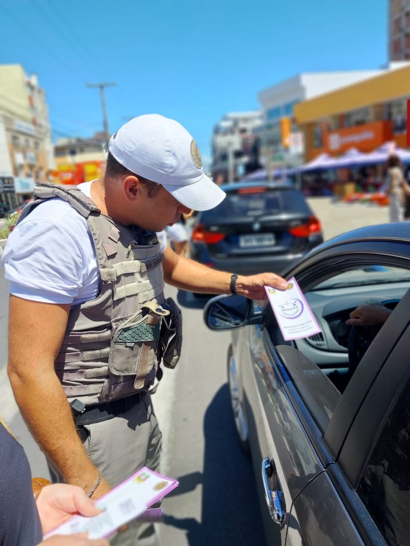 Brigadiano está fardado entregando um panfleto para o motorista de um carro preto.