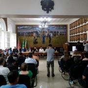 Grupo de pessoas sentadas está reunido em uma sala, voltado em direção a uma pintura. 