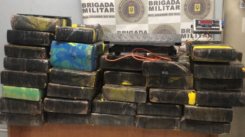 Foto mostra diversos tijolos de maconha empilhados em cima de uma mesa, junto a uma balança. Por trás, o banner da BM