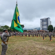 Foto mostra soldado segurando bandeira do Brasil, em campo aberto, em frente aos formandos perfilados