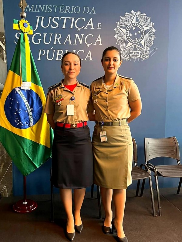Duas militares estão lado a lado olhando para a câmera ao lado de uma bandeira do Brasil