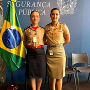 Duas militares estão lado a lado olhando para a câmera ao lado de uma bandeira do Brasil