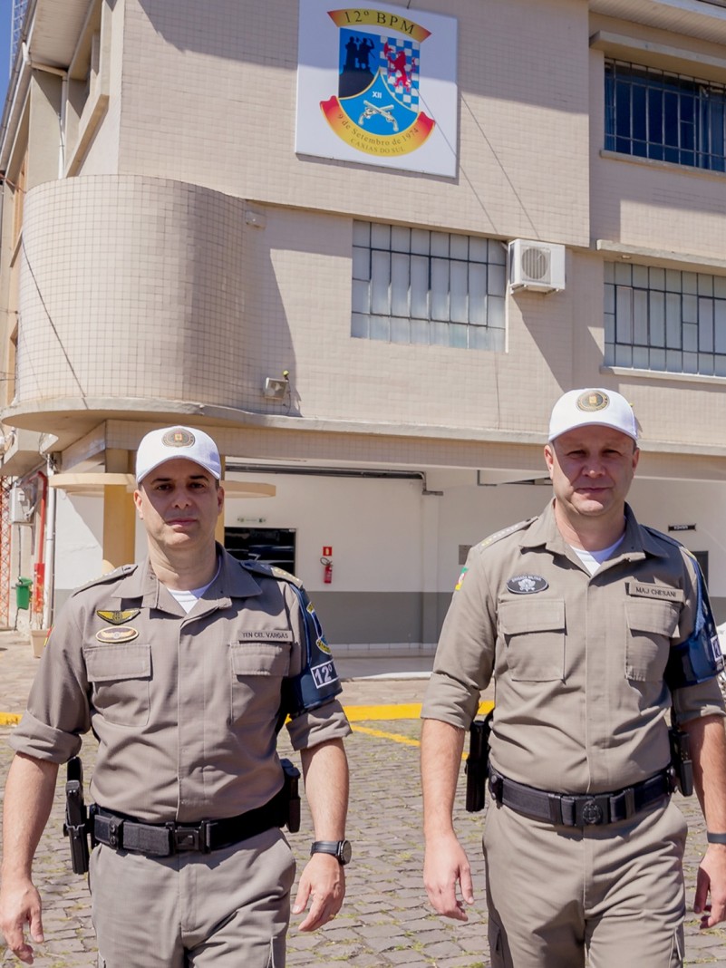 Dois militares estão caminhando em direção a câmera, em frente ao prédio do 12° batalhão.