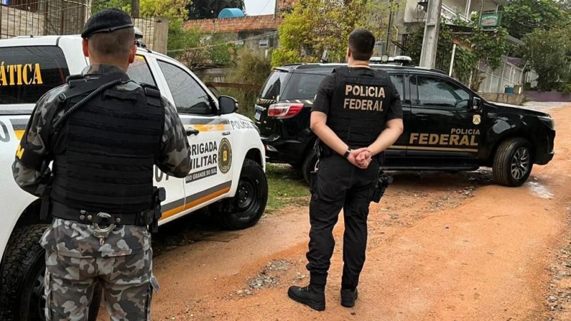 Foto mostra um policial militar e um agente da Polícia Federal em uma rua de barro. Ambos estão de costas, em frente a duas viaturas que estão estacionadas em frente a uma casa.
