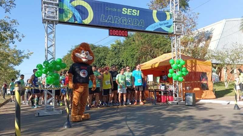 O boneco em tamanho humano de um leão que é mascote do Proerd, junto aos participantes da corrida do SESI na linha de largada