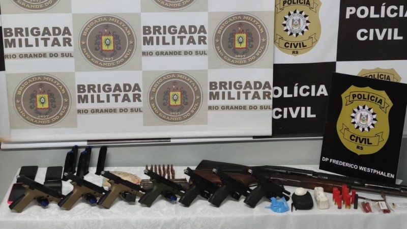 Imagem mostra armas de fogo dispostas em cima de uma mesa. Por trás, os banners da Brigada Militar e da Polícia Civil.