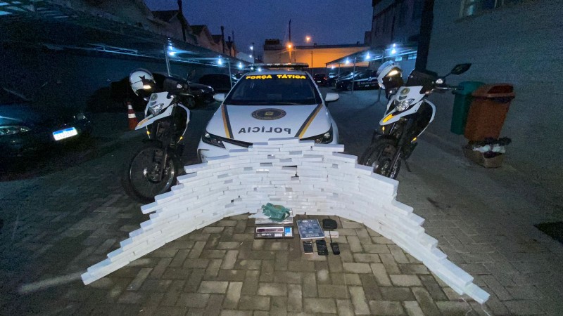 Materiais apreendidos estão empilhados em frente a uma viatura e duas motos da BM