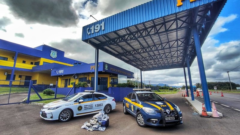 Foto mostra duas viaturas, sendo uma da BM e outra da PRF, paradas em um posto da PRF. Entre elas, está uma mala aberta com embalagens de maconha